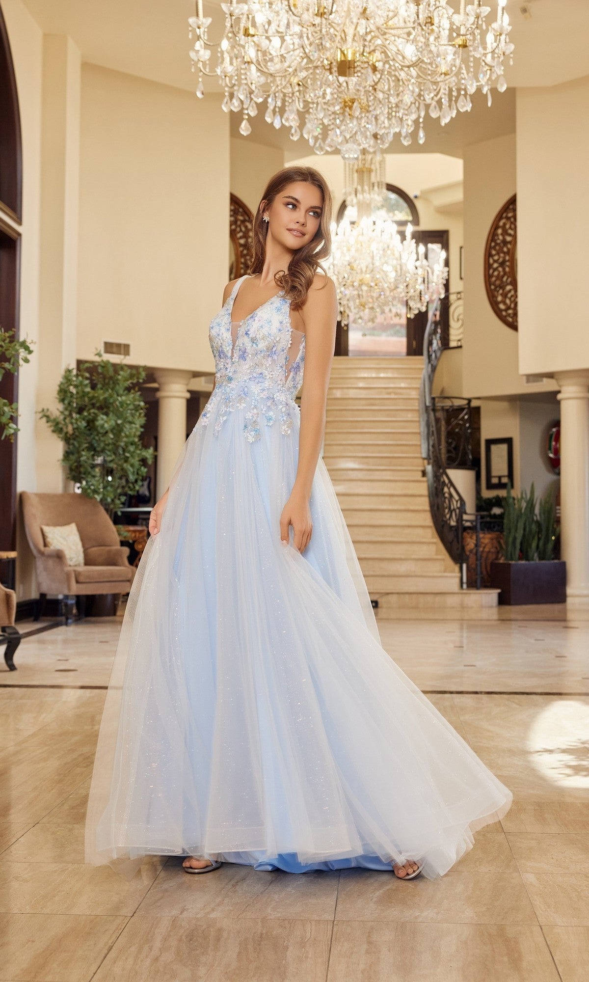 Wearing the Nox Anabel Y1799 long prom dress with pastel tulle and delicate floral details, a woman stands in an elegant foyer beneath sparkling chandeliers, framed by a grand staircase.